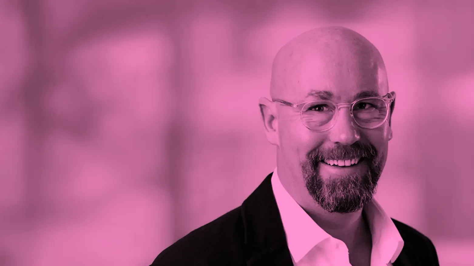 Porträtfoto eines lächelnden, glatzköpfigen Mannes mit runder Brille und kurzem Bart in dunklem Sakko über hellem Hemd, das gesamte Bild in Pink-Magenta-Tönen gehalten.