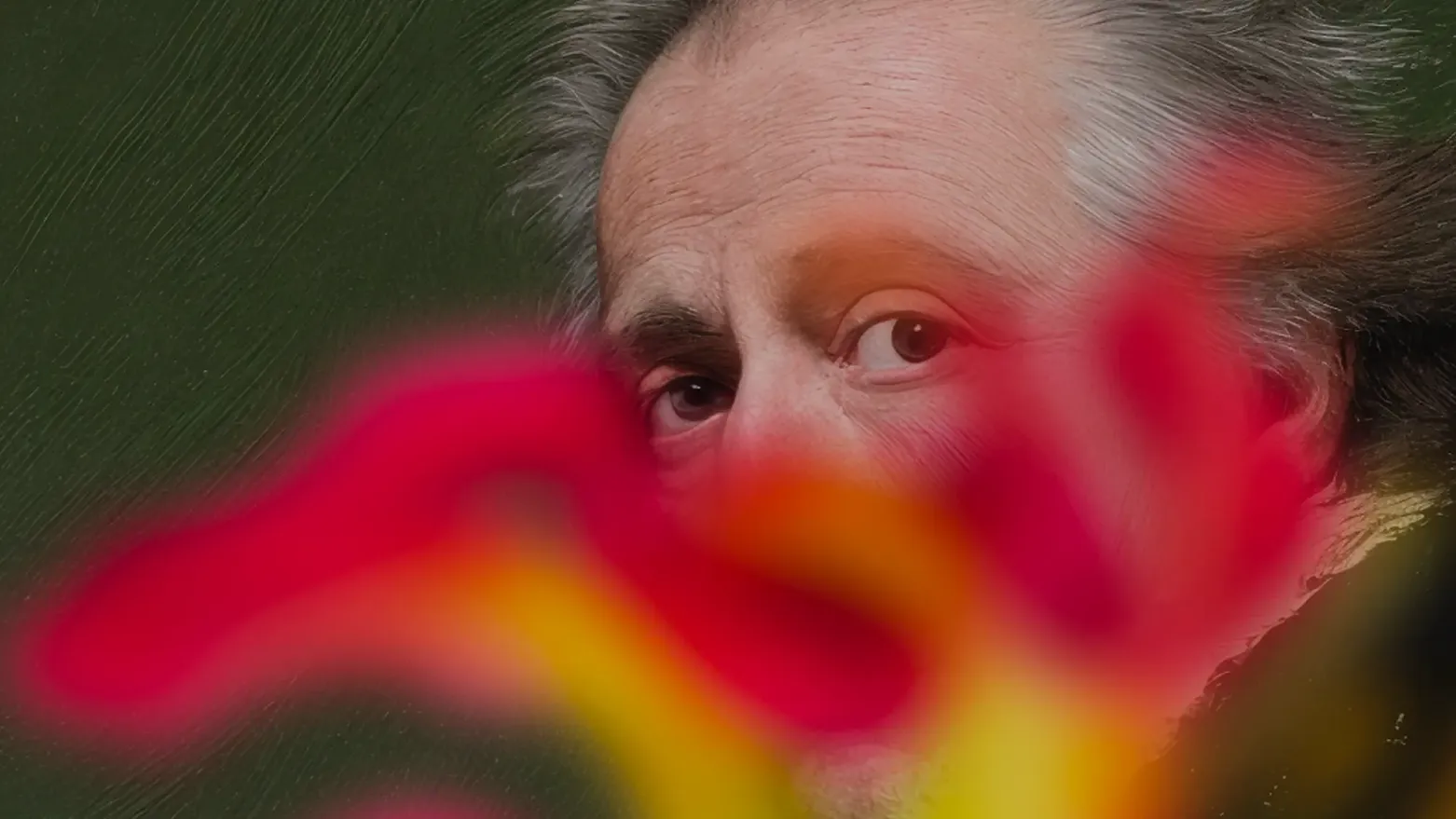 Nahaufnahme des Gesichts eines älteren Mannes mit grauem Haar, dessen untere Gesichtshälfte von einer unscharfen roten und gelben Blüte im Vordergrund verdeckt wird, vor dunkelgrünem Hintergrund.